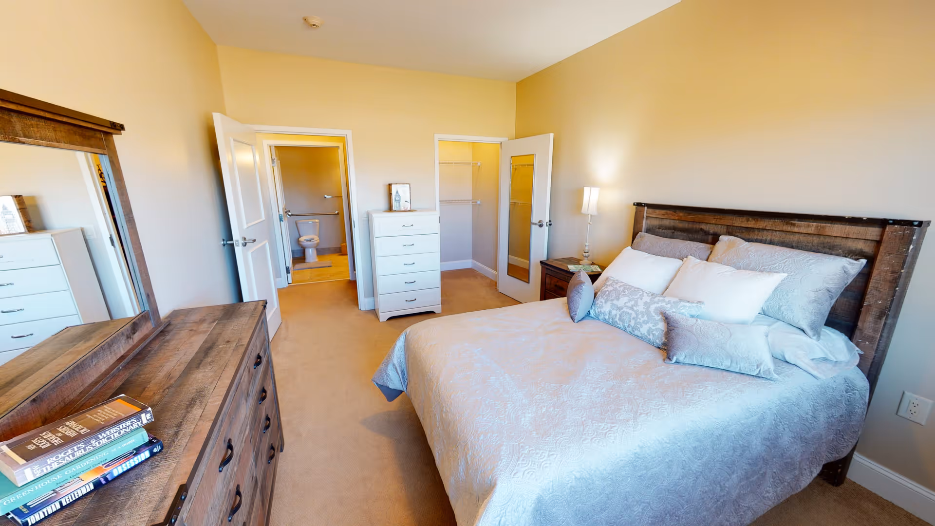 Bright furnished bedroom with a bed, wooden dresser and mirror, nightstand, and a view into an en-suite bathroom.