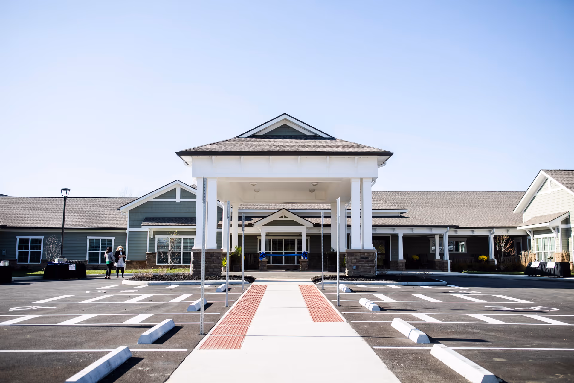 Front exterior view of the Laurels of West Columbus facility showing a covered entrance with white pillars, a wide sidewalk with red tactile paving, and an empty parking lot under a clear blue sky.