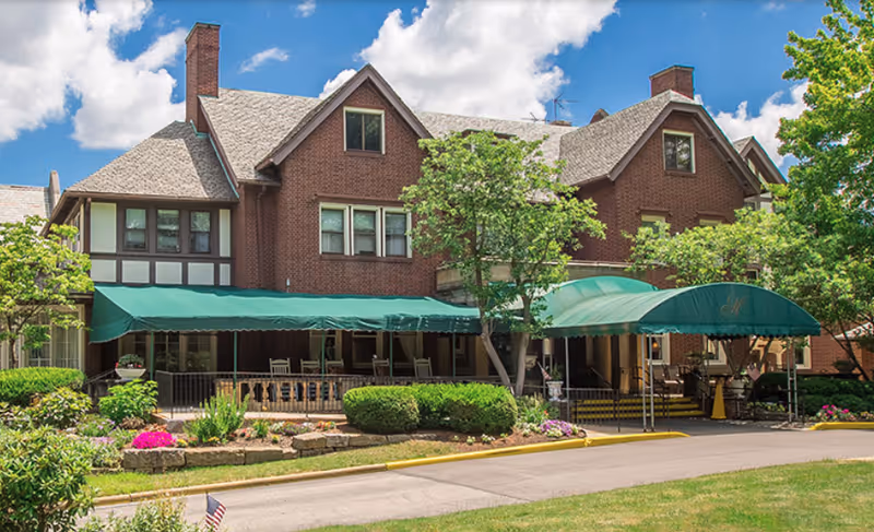 Brick multi-story building front with green fabric awnings over a porch and entrance surrounded by landscaped shrubs and flowers.
