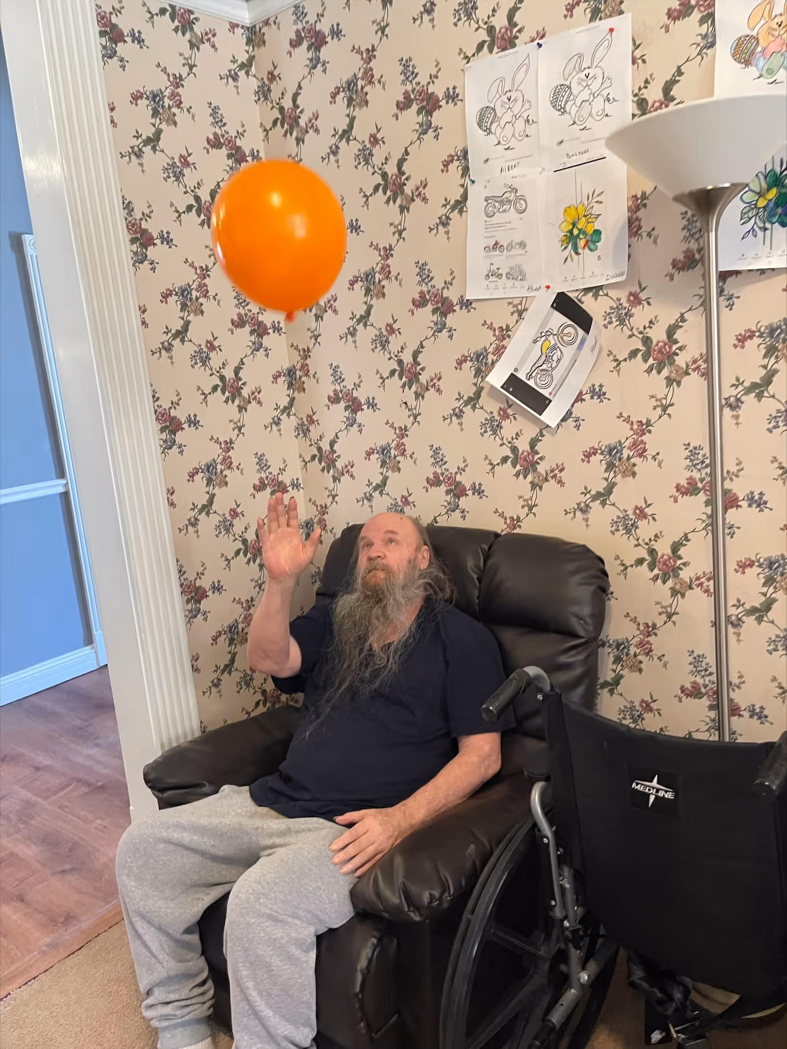 An elderly man with a long beard sitting in a brown recliner chair in a room with floral wallpaper. He is reaching up towards an orange balloon floating above him. A wheelchair is positioned next to the chair. Several coloring pages with drawings of bunnies and flowers are taped to the wall behind him, along with a tall floor lamp.