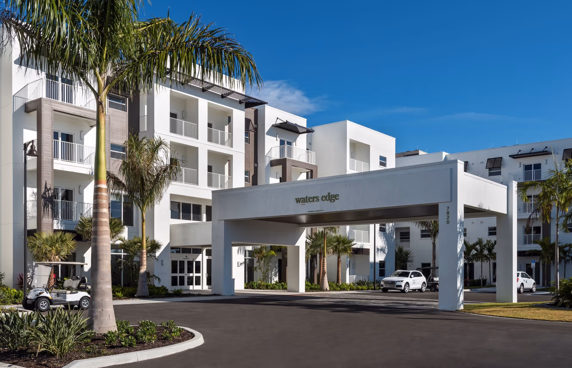 Exterior view of a modern multi-story senior living facility named Waters Edge with white walls, balconies, palm trees, and a covered driveway entrance. Several cars and golf carts are parked near the entrance under a clear blue sky.