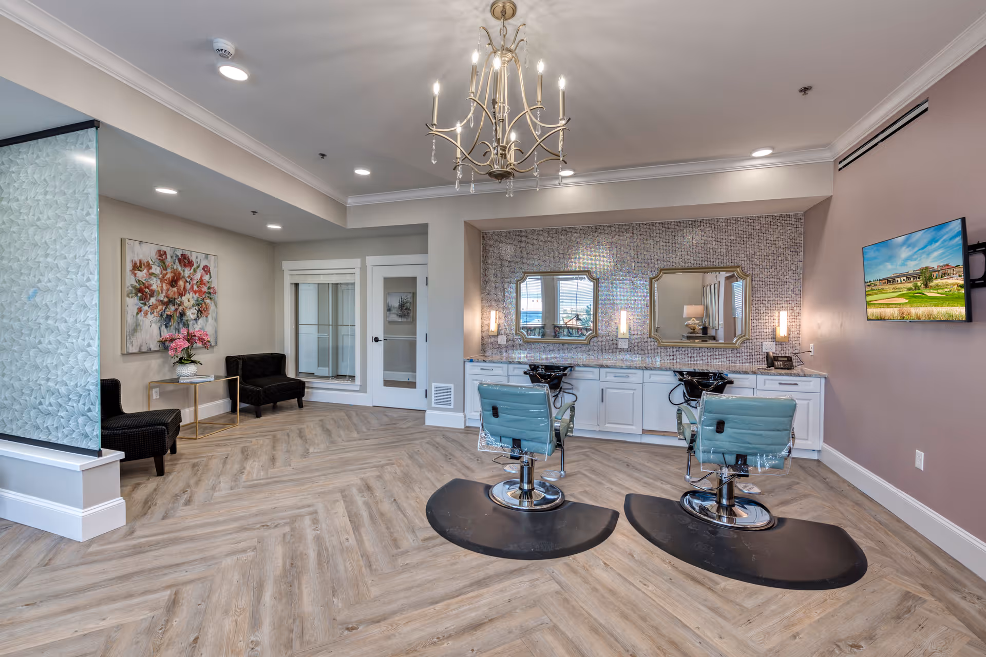 A modern salon area in a senior living facility featuring two styling chairs in front of a counter with two mirrors and sinks. The room has wood-patterned flooring, a chandelier, a wall-mounted TV, and a seating area with two black chairs and a small table with flowers. The walls are decorated with a floral painting and a textured accent wall behind the mirrors.
