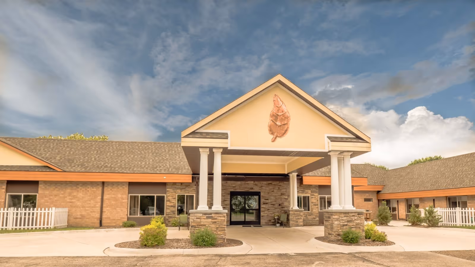 Front entrance of a single-story brick senior living building with a covered porte-cochere supported by columns under a partly cloudy sky.