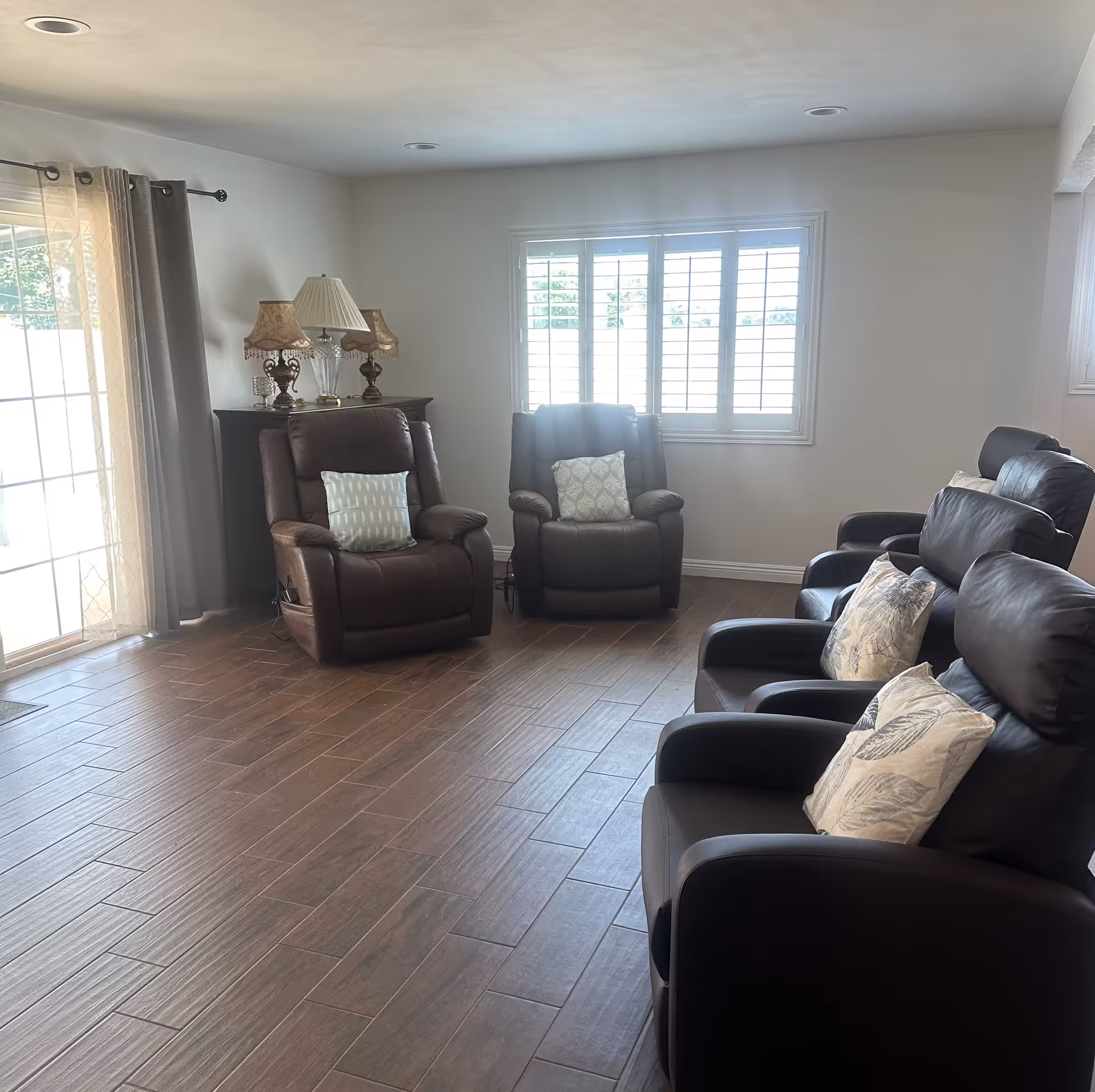 Sunlit living room with several brown leather recliners, decorative pillows, a side table with lamps, and a window with shutters.