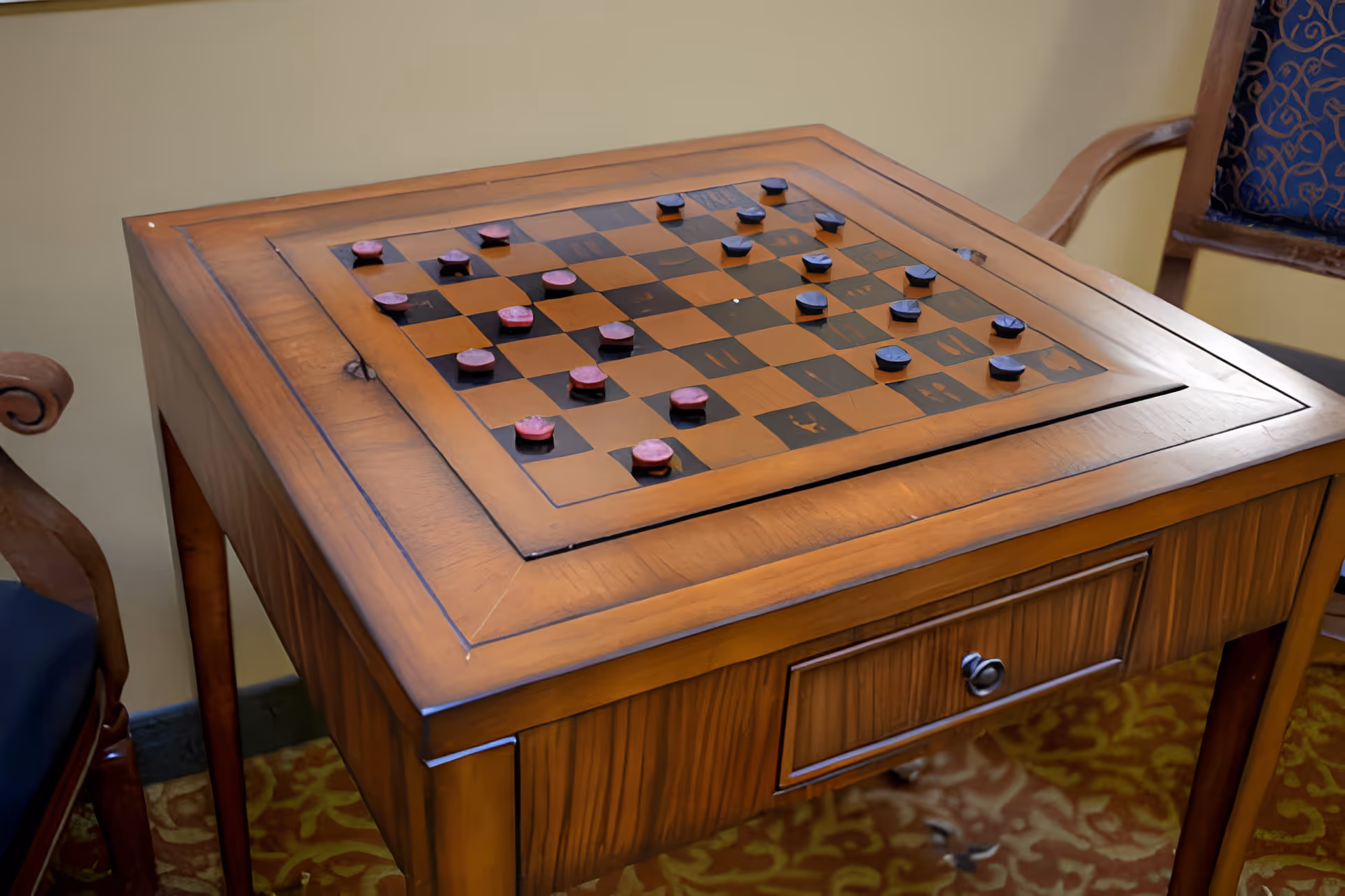 A wooden table with an integrated checkers board and checkers pieces set up for a game, surrounded by wooden chairs with blue patterned cushions, placed on a patterned carpet.