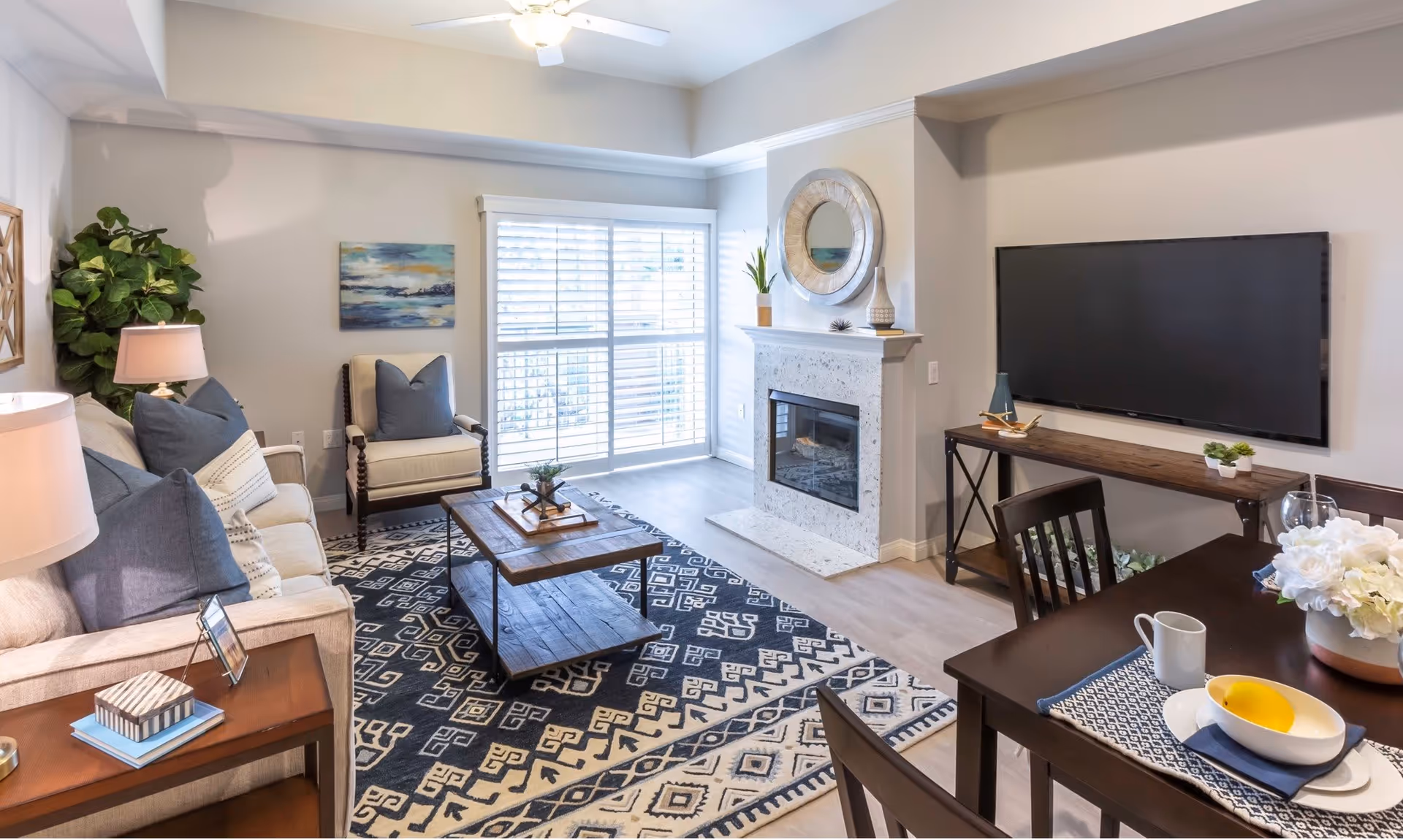 A bright and cozy living room with a beige sofa adorned with blue and white pillows, a wooden coffee table on a patterned rug, a beige armchair, a fireplace with decorative items on the mantel, a large flat-screen TV mounted on the wall, and a dining table set with white flowers, a bowl with a yellow fruit, and a cup. The room has large windows with white blinds allowing natural light to fill the space.
