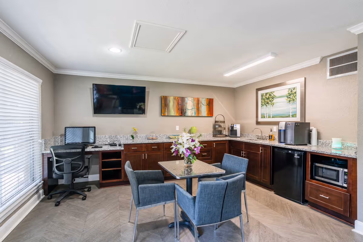 A bright and modern common area with a small dining table surrounded by four blue upholstered chairs. The room features granite countertops with dark wooden cabinets, a mini refrigerator, microwave, coffee machine, and a computer workstation with an office chair. Two framed artworks hang on the walls, and a flat-screen TV is mounted above the countertop. Large window blinds allow natural light into the space.