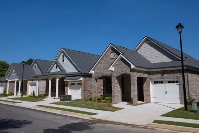 A row of single-story brick houses with garages, driveways, and small front lawns under a clear blue sky, with a streetlamp visible on the right side.