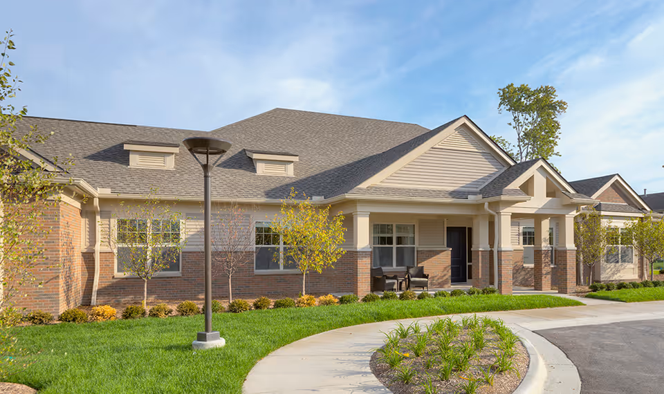 Exterior view of a single-story senior living facility building with brick and siding facade, a covered entrance with columns, a curved sidewalk, green lawn, small trees, and a streetlamp under a blue sky.