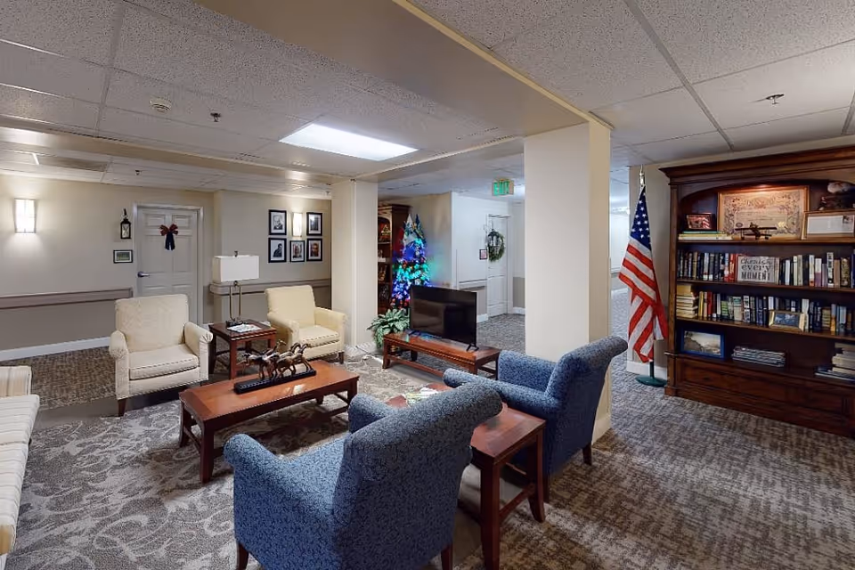 A cozy senior living facility common area with comfortable armchairs and sofas arranged around a wooden coffee table. There is a small TV on a stand, a decorated Christmas tree, an American flag, and a large wooden bookshelf filled with books and decorative items. The room has carpeted floors, beige walls, and ceiling lights providing bright illumination.