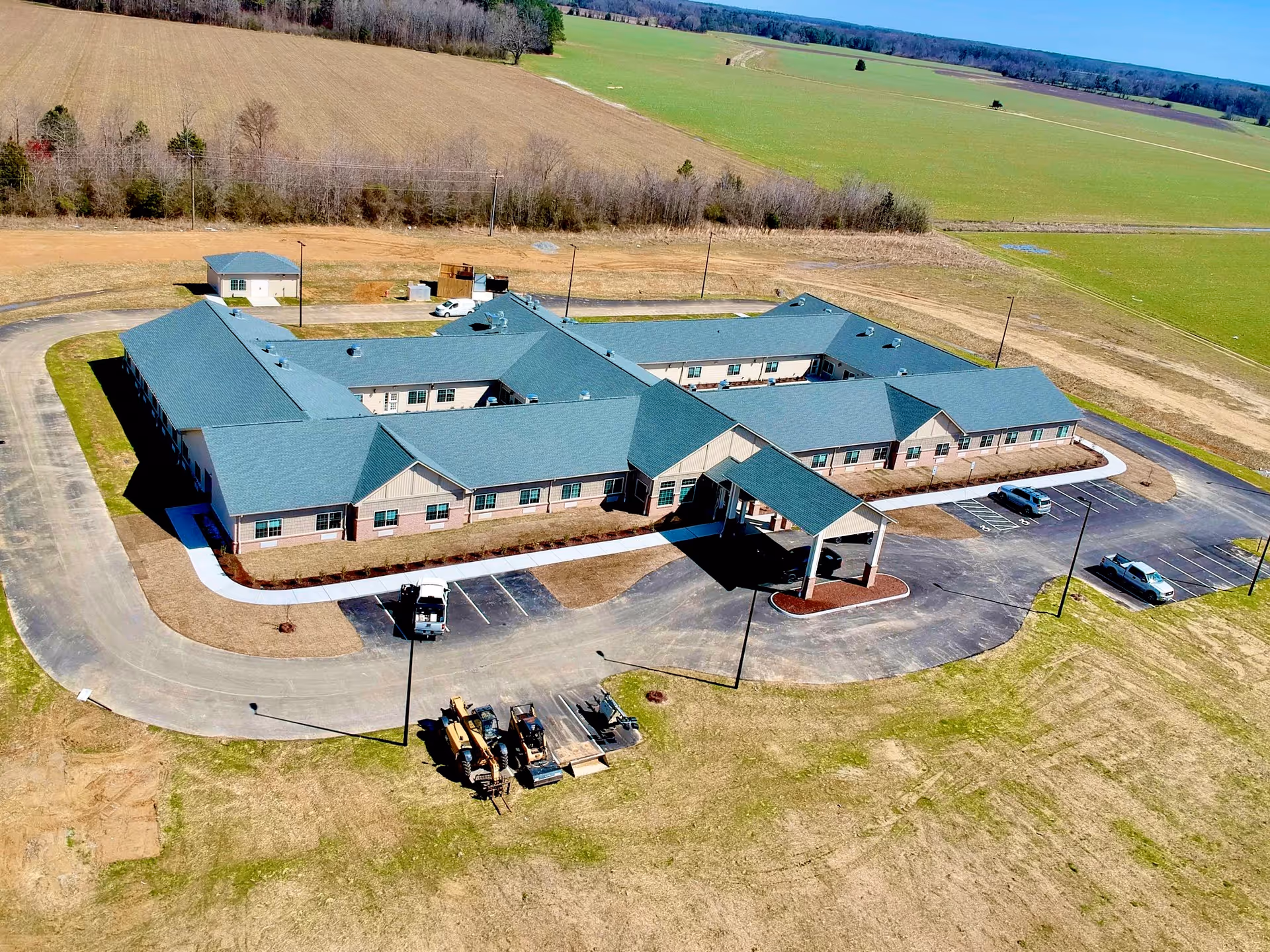 Aerial view of a single-story, U-shaped senior living building with a porte-cochere, parking lot, and surrounding farmland.
