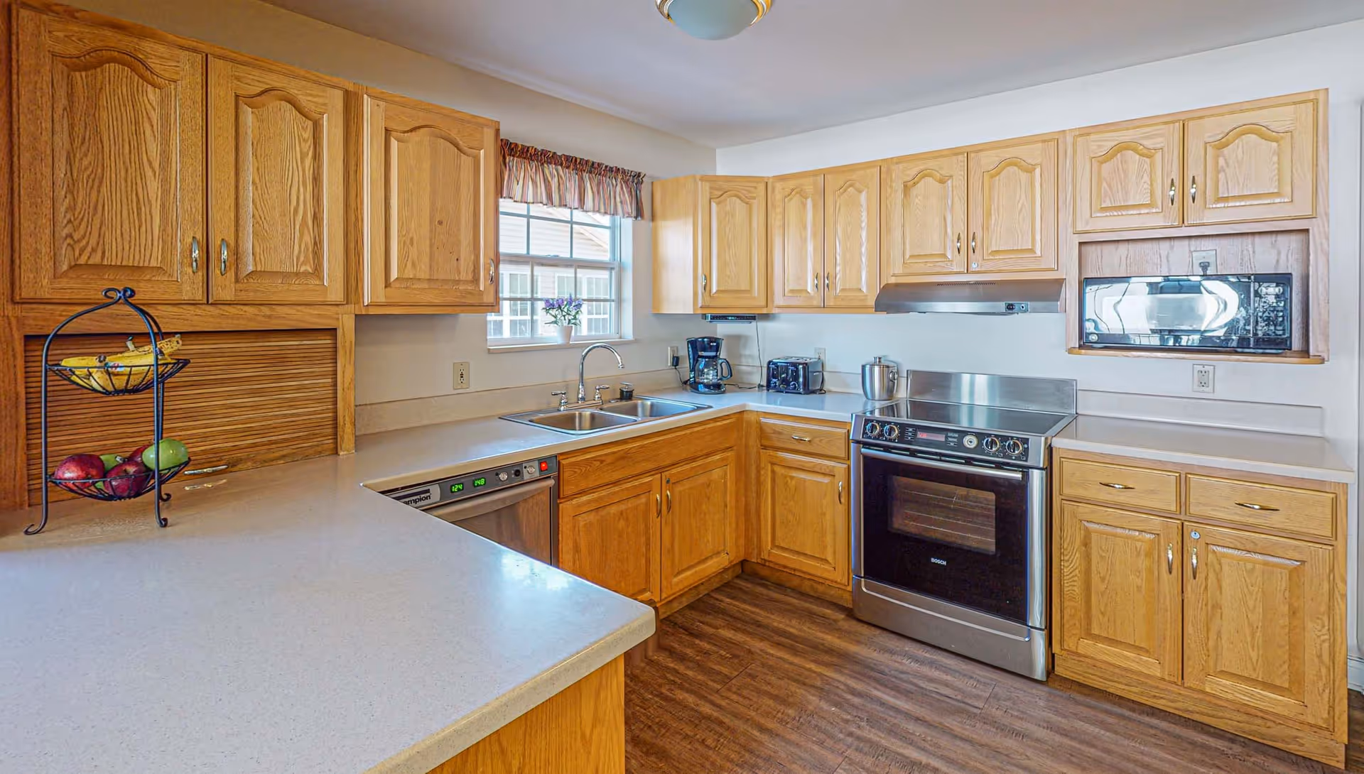 A bright kitchen with wooden cabinets, a stainless steel oven and microwave, a double sink under a window with a small flower pot, and a countertop with a two-tier fruit basket containing bananas, apples, and pears.