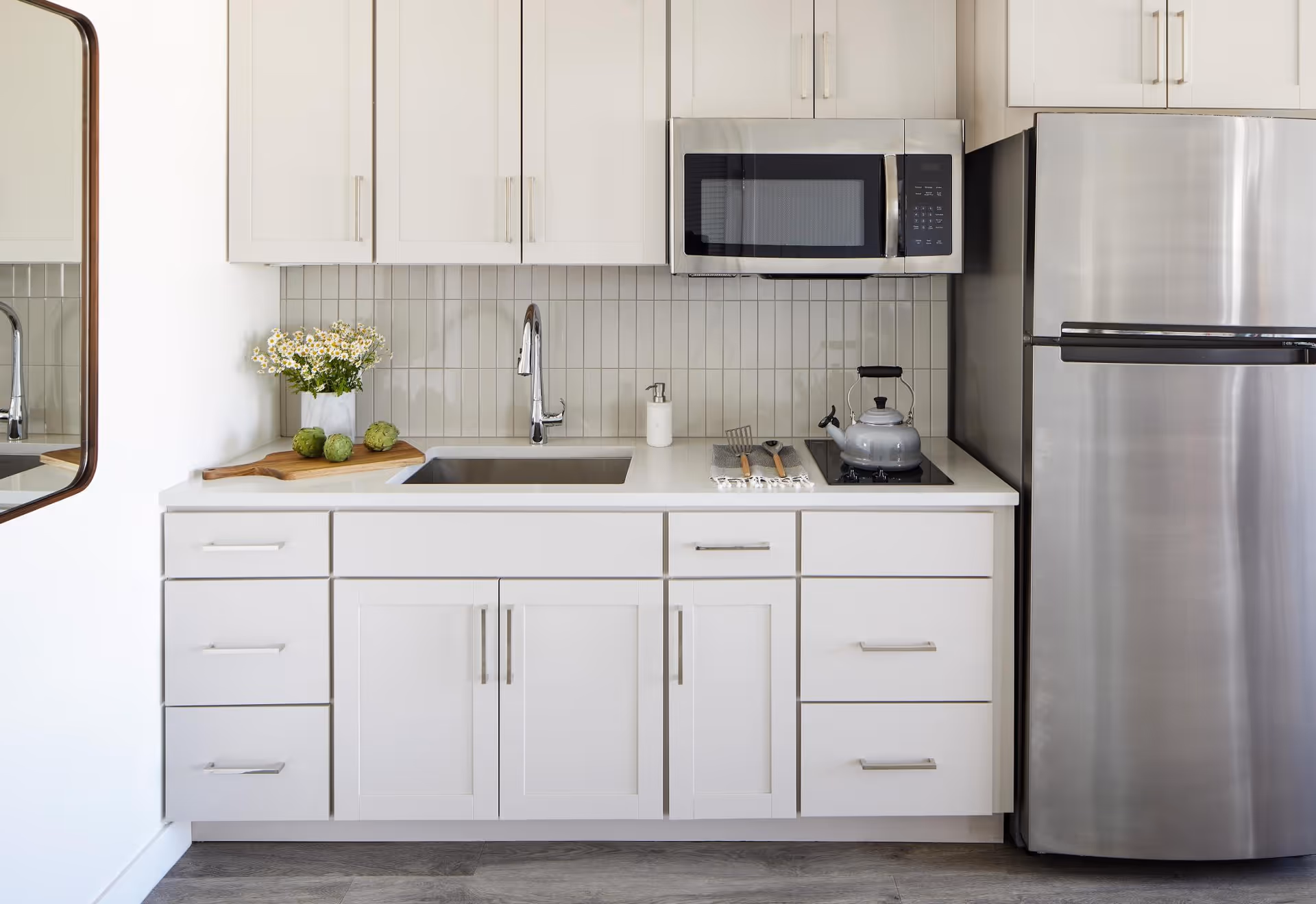 A modern kitchen with white cabinets and drawers, a stainless steel refrigerator, a built-in microwave above a stovetop with a kettle, a sink with a faucet, and a countertop decorated with a vase of flowers and some artichokes on a wooden cutting board.