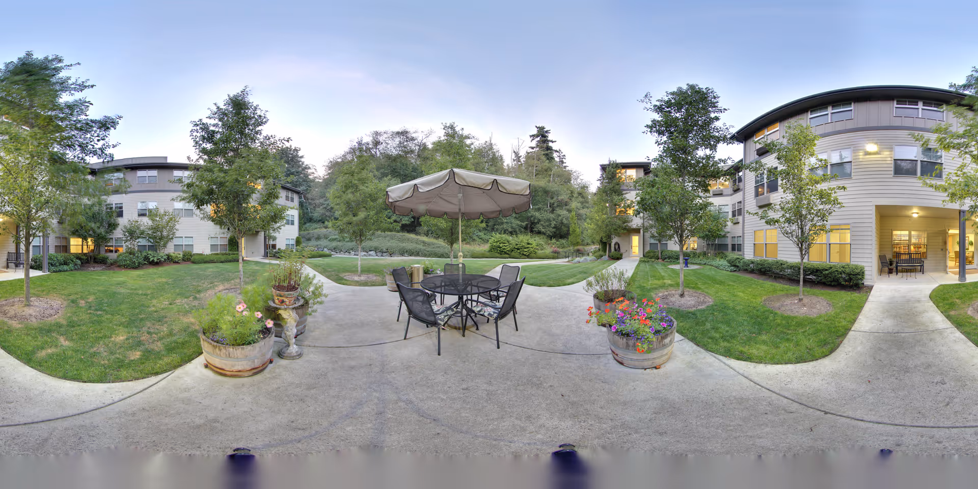 Outdoor patio area at Maple Ridge by Bonaventure featuring a round table with six chairs and a large umbrella. The patio is surrounded by green grass, trees, and flower pots with colorful flowers. In the background, there are multi-story residential buildings with lit windows and a pathway leading through the garden area.