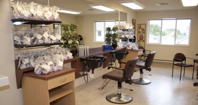 Interior view of a senior living facility hair salon with two salon chairs and hair washing stations. Shelves hold numerous plastic bags with hair care supplies. Large windows let in natural light, and there are a few chairs along the wall for waiting.