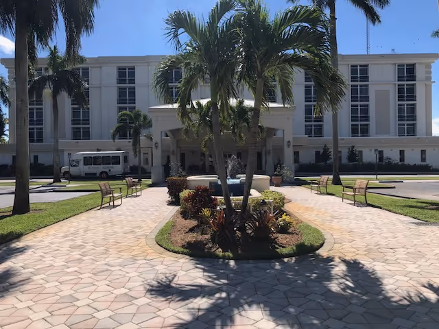 Front exterior view of The Peninsula Assisted Living & Memory Care building with a paved walkway, palm trees, benches, and a fountain in the center under a clear blue sky.