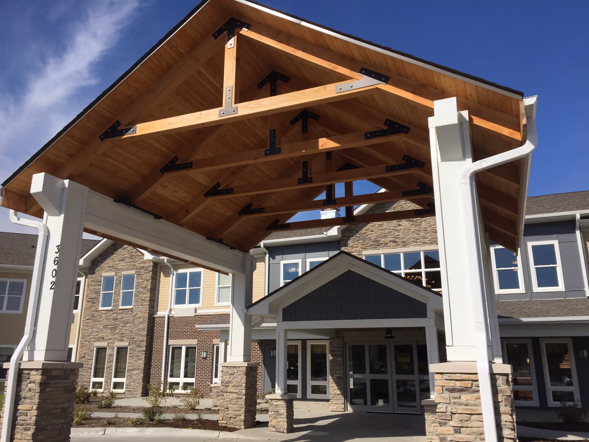 Entrance of a senior living facility with a large wooden canopy supported by white pillars with stone bases. The building has multiple windows and a combination of stone and siding exterior under a clear blue sky.