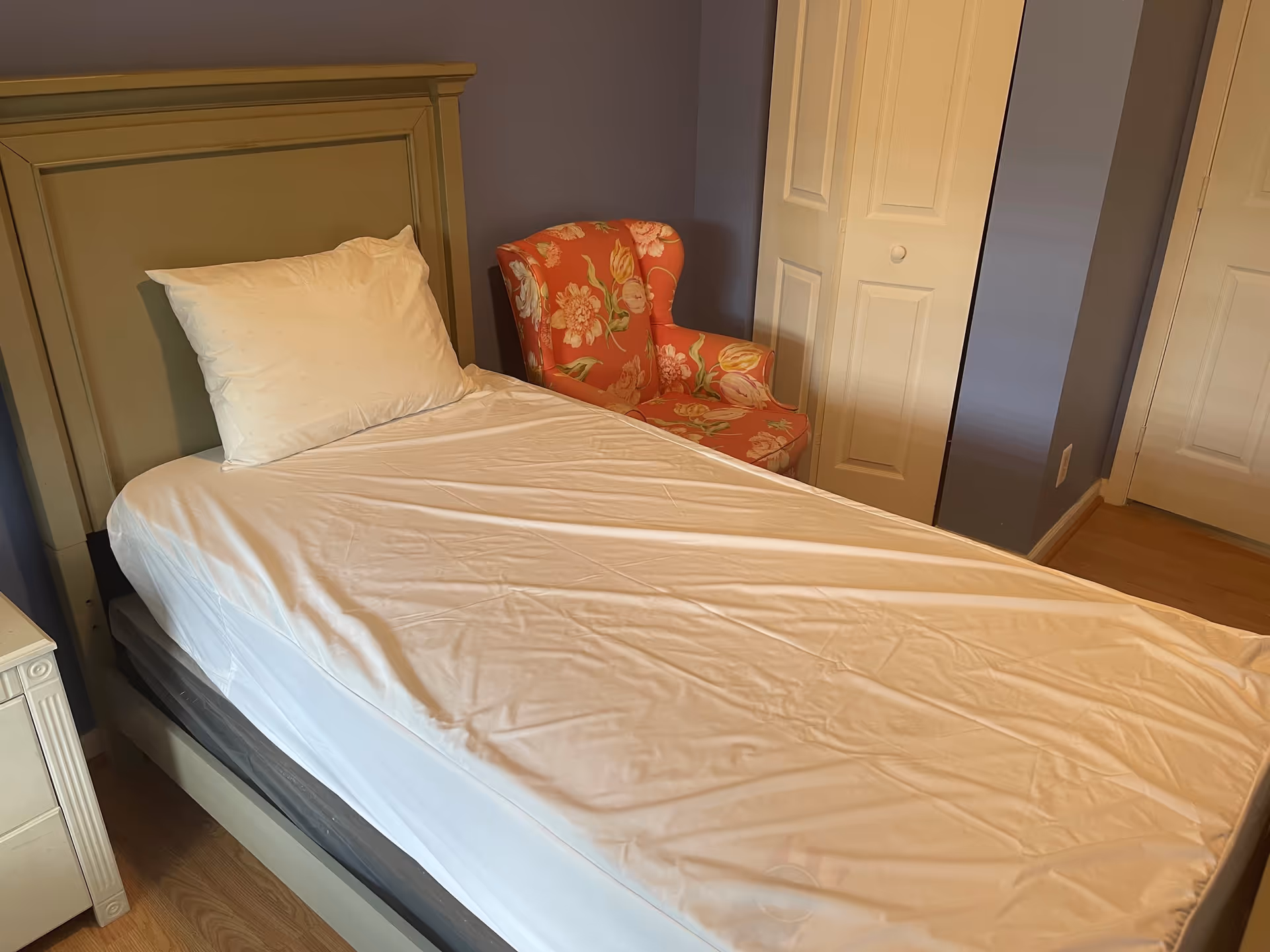 A bedroom with a single bed featuring a green headboard and white pillow and sheets. Next to the bed is a floral patterned armchair in shades of orange and pink. The room has light wooden flooring, a white nightstand, and a white closet door against a purple wall.
