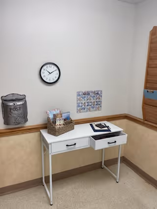 Small white desk in a beige corner with a wall clock, a basket of mail, an open drawer, and wall decorations.