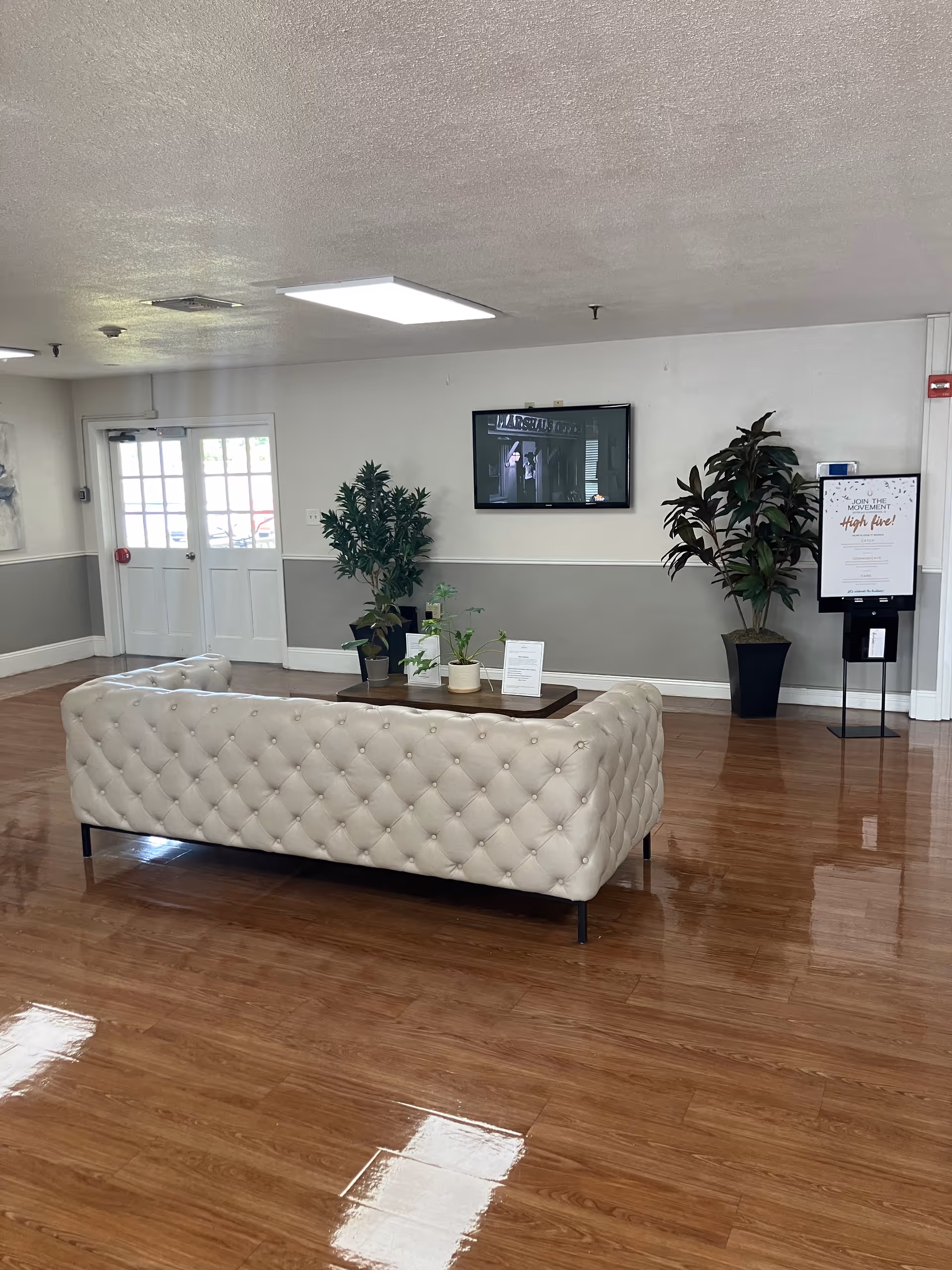 Bright reception lounge with a tufted white sofa, coffee table, potted plants, wall-mounted TV, and an entry door.