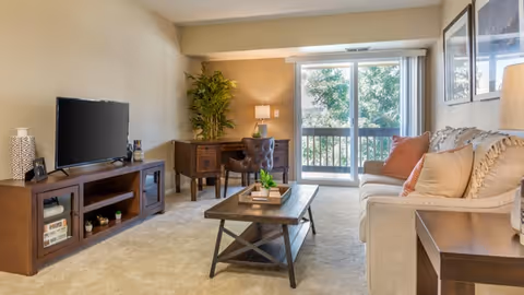 A cozy living room with a beige sofa adorned with multiple pillows, a wooden coffee table with a small plant, a wooden TV stand with a flat-screen TV, and a desk with a chair and lamp in the corner. Large sliding glass doors lead to a balcony with greenery visible outside.