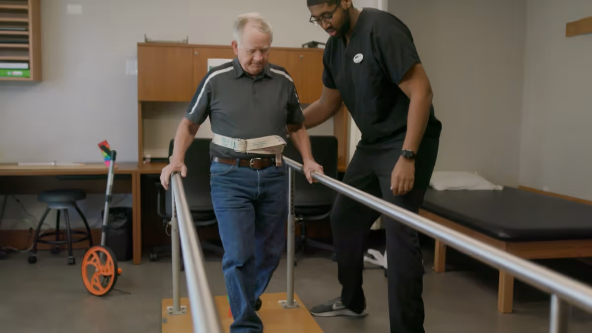 An elderly man is walking between parallel bars for physical therapy assistance, supported by a healthcare professional in a clinical room with a desk, chair, and examination table in the background.