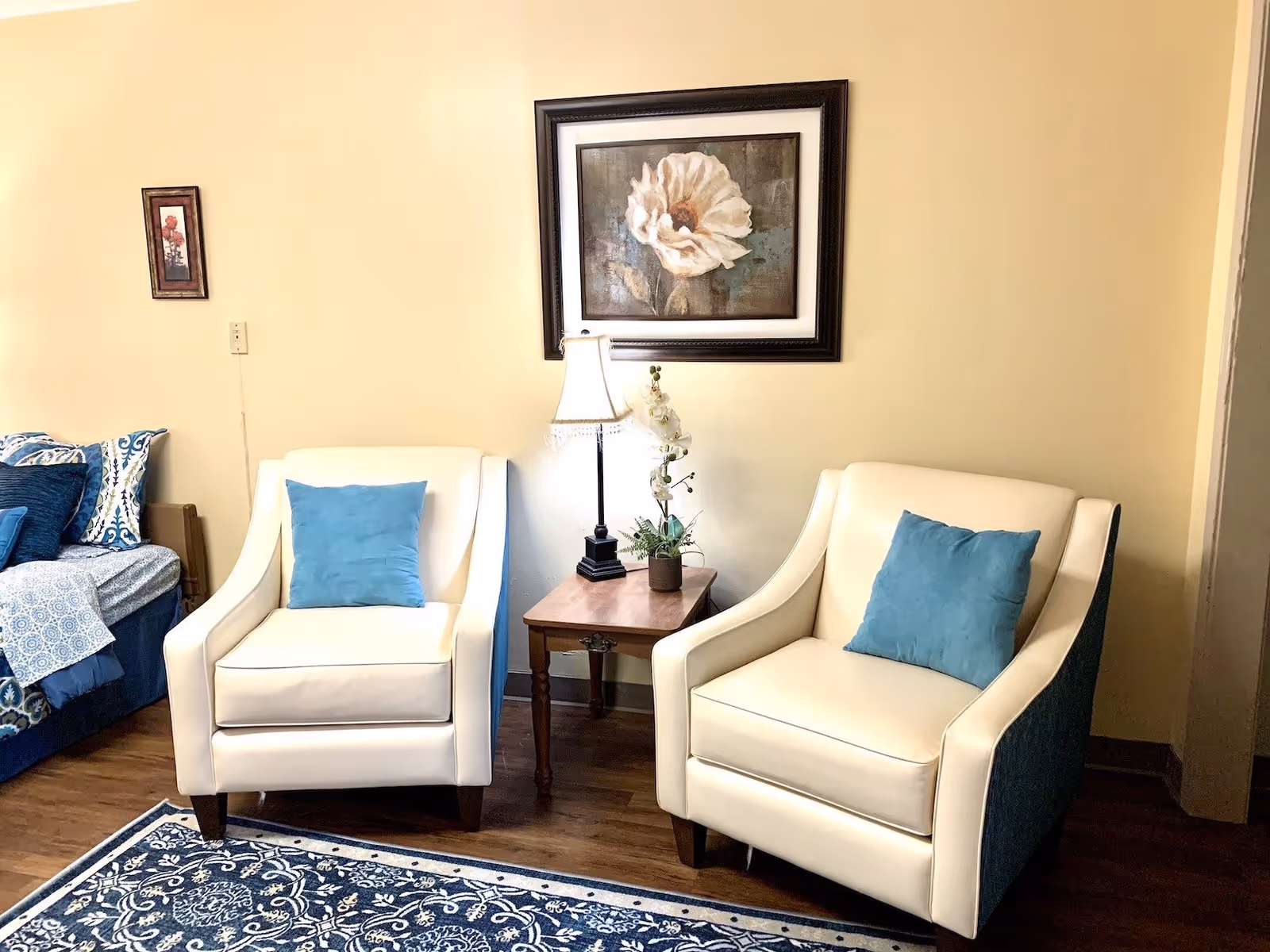 A cozy sitting area with two cream-colored armchairs each with a blue cushion. Between the chairs is a wooden side table with a lamp and a small potted plant. Above the table hangs a framed painting of a white flower. To the left, part of a bed with blue and white bedding is visible. The room has light yellow walls and a blue patterned rug on a wooden floor.