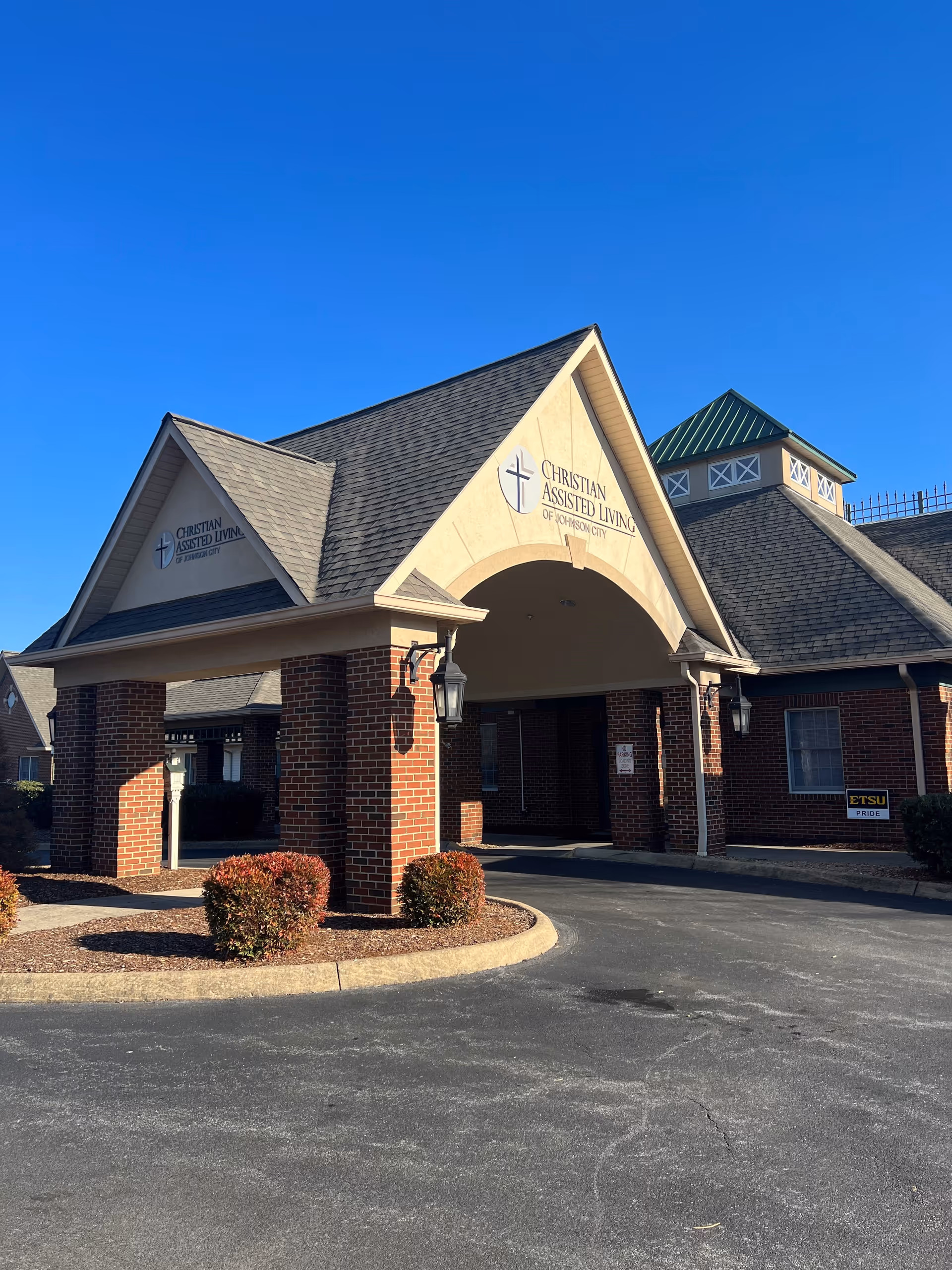 Exterior view of the entrance to Christian Assisted Living of Johnson City, featuring a covered driveway supported by brick columns, a peaked roof, and clear blue sky above.