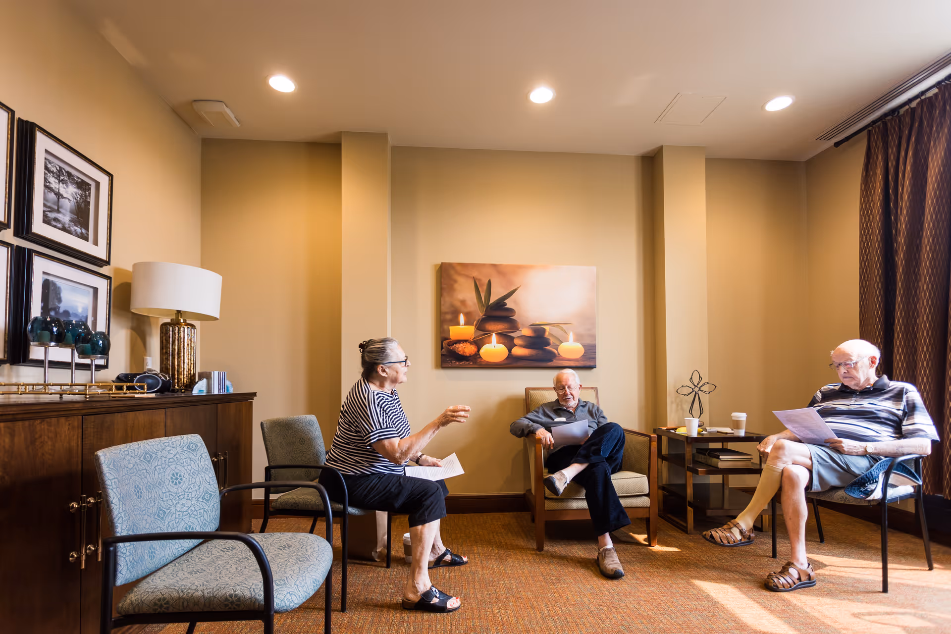 Three elderly people sitting in a cozy room with beige walls and carpeted floor. Two men and one woman are seated on chairs arranged in a semi-circle, holding papers and engaging in conversation. The room has framed pictures on the wall, a lamp on a wooden cabinet, and a painting of candles and stones on the wall. A small table with cups and decorative items is also present.