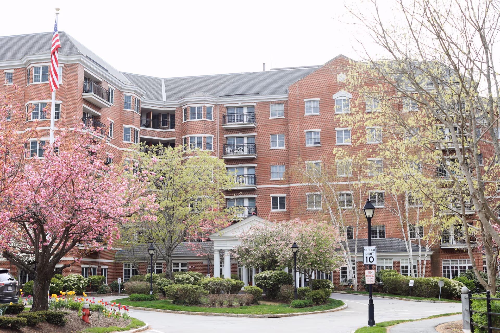 Exterior view of a multi-story brick building with balconies, surrounded by blooming trees and landscaped greenery. An American flag is visible on a flagpole to the left, and a driveway with a speed limit sign is in front of the building.
