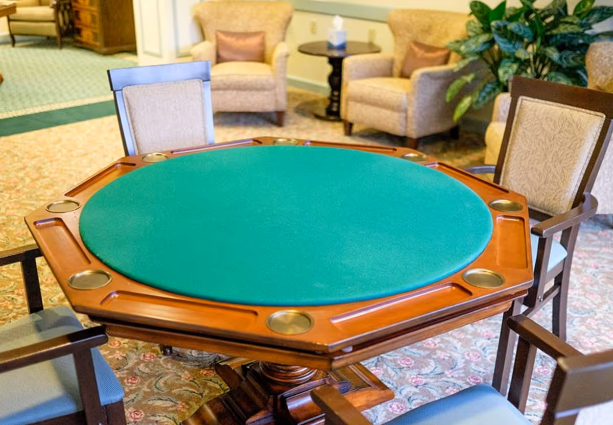 A cozy seating area with a green felt octagonal game table surrounded by four wooden chairs with cushioned seats. In the background, there are two beige upholstered armchairs with brown accent pillows, a small round wooden side table with a tissue box, and a large leafy plant. The room has a patterned carpet and soft lighting.