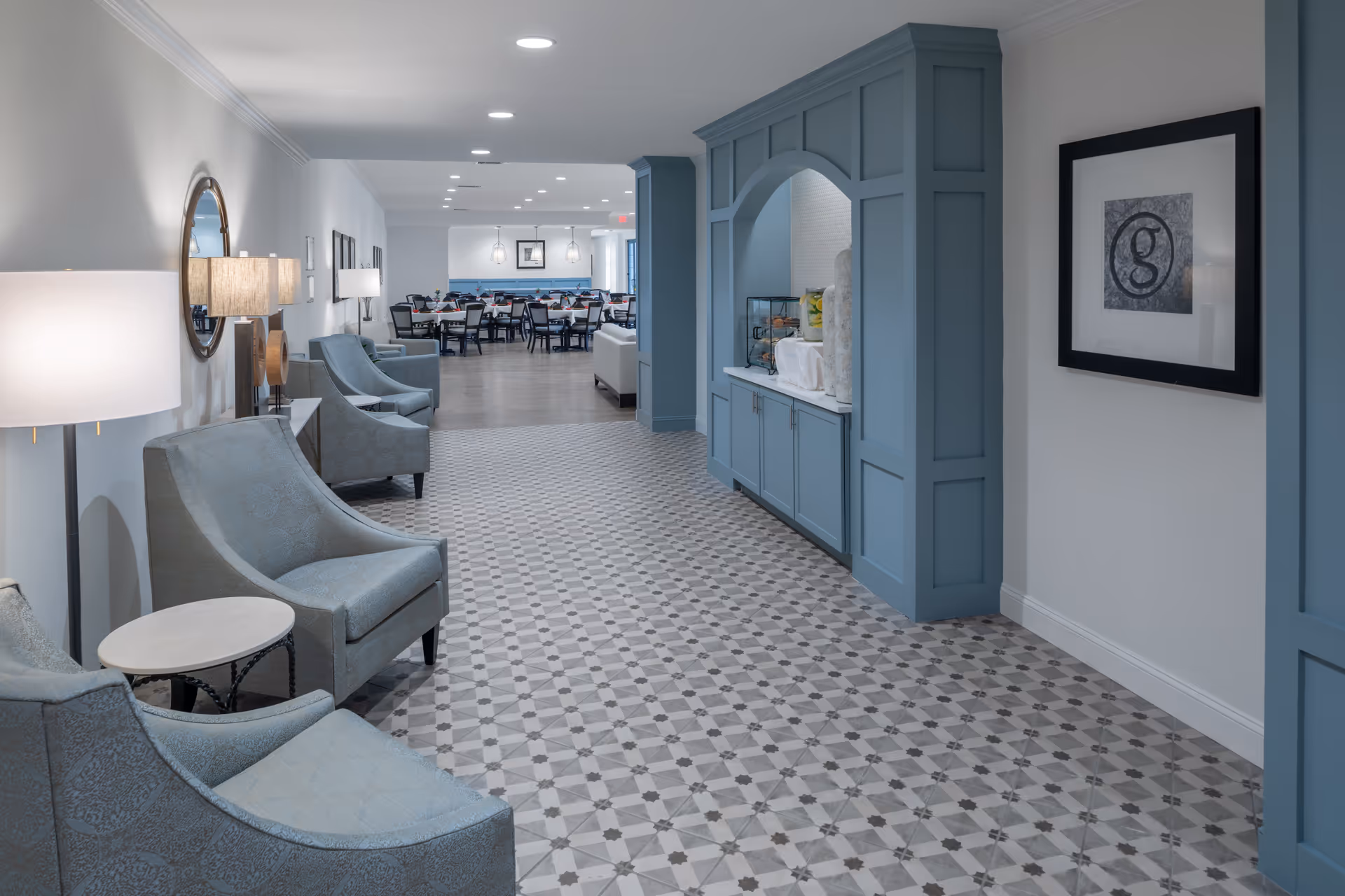 A spacious, well-lit interior hallway with patterned tile flooring, light blue armchairs and side tables along the left wall, and a built-in blue cabinetry unit on the right. The hallway leads to a dining area with multiple tables and chairs in the background. The walls are painted white with decorative elements including a round mirror and framed artwork.