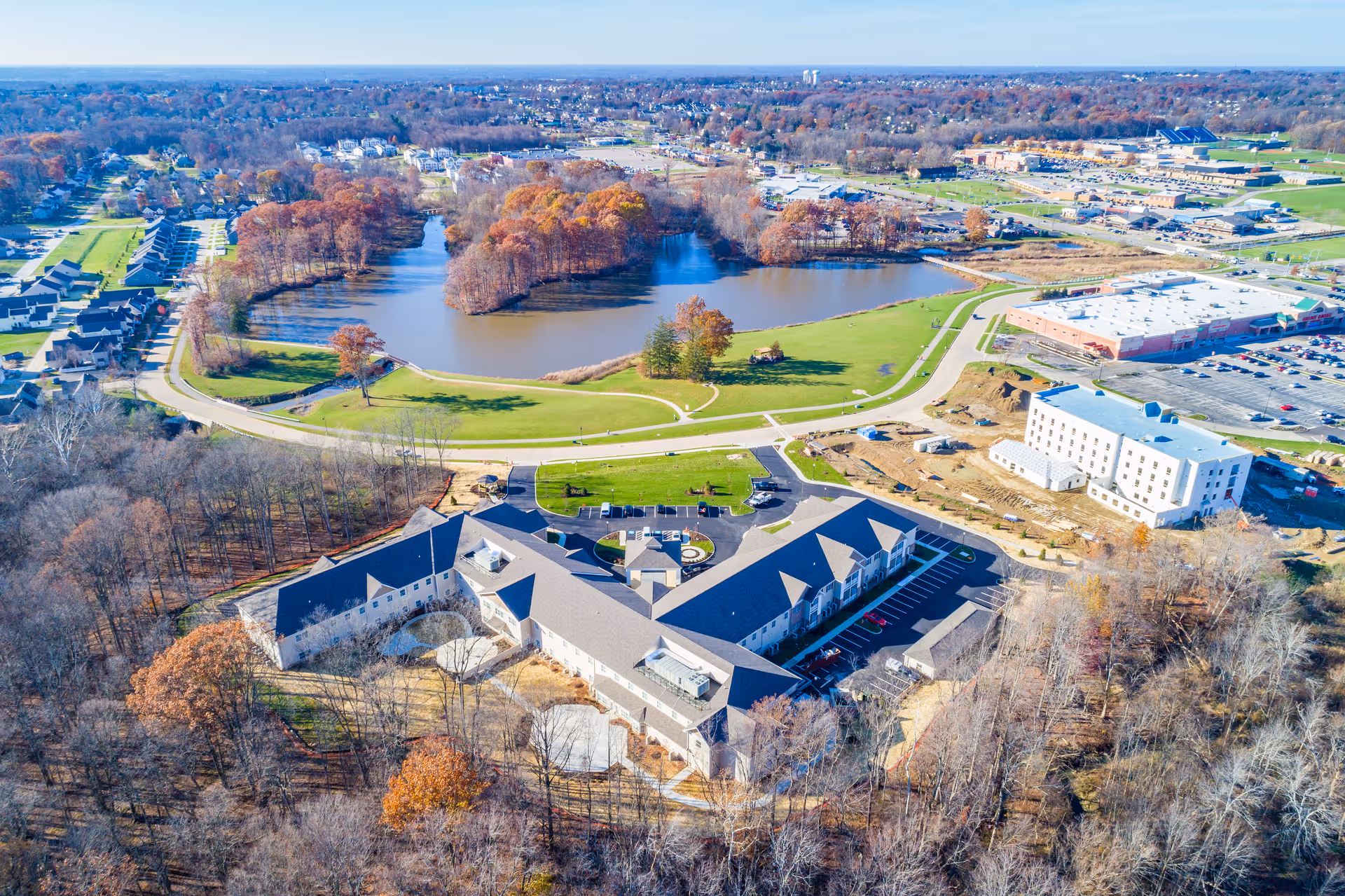 Aerial view of a senior living facility named Danbury Brunswick surrounded by trees with autumn foliage, a large pond with small islands, nearby residential houses, and commercial buildings. The facility has a large, U-shaped building with parking lots and green spaces around it.