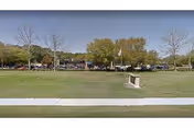 Wide grassy lawn with a sidewalk in the foreground and a row of trees and parked cars in the background under a clear sky.