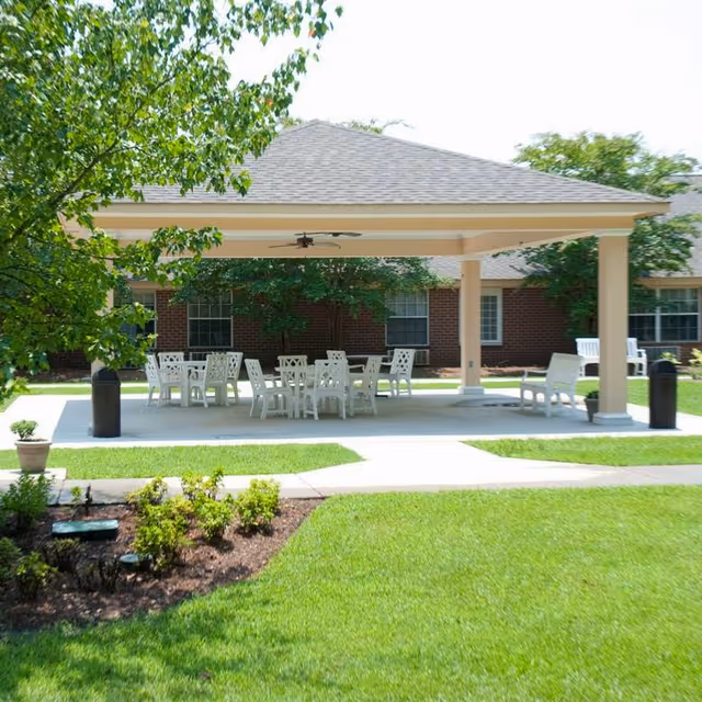 Outdoor covered patio area with white tables and chairs set on a concrete slab surrounded by green grass and landscaping. The patio has a ceiling fan and is located in front of a brick building with windows.