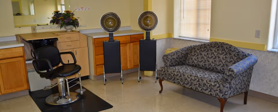 Interior room with a black salon chair on a black mat, two vintage hair dryers on stands, wooden cabinets with a countertop, a floral arrangement in a vase, and a patterned upholstered loveseat near a window with blinds.