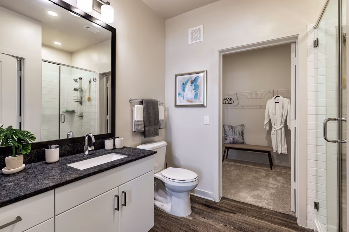 Modern bathroom with a large mirror above a black granite countertop with a sink. There is a white toilet next to the counter, and a towel rack with folded towels above it. A glass-enclosed shower with white subway tiles is visible. An open doorway leads to a carpeted walk-in closet with a bench, a pillow, hangers, and a white robe hanging on the wall.
