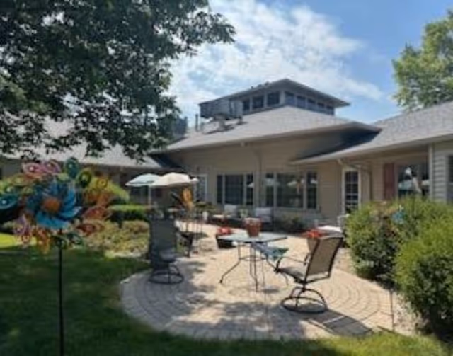 Outdoor patio area at Courtyard Estates of Monmouth with a round paved seating area featuring metal chairs and tables, surrounded by greenery and bushes under a partly cloudy sky.