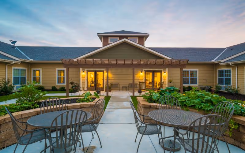 Outdoor courtyard with metal patio tables and chairs, raised planters and a pergola leading to the lit entrance of a single-story senior living building.