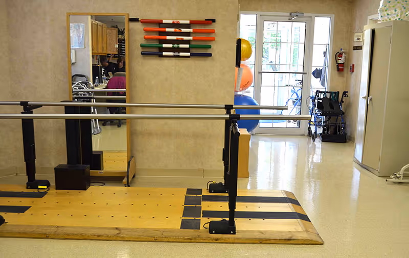 Physical therapy room with parallel bars for walking exercises, a large mirror on the wall, colorful exercise sticks mounted above the mirror, exercise balls near a glass door, and a walker and wheelchair near a cabinet.