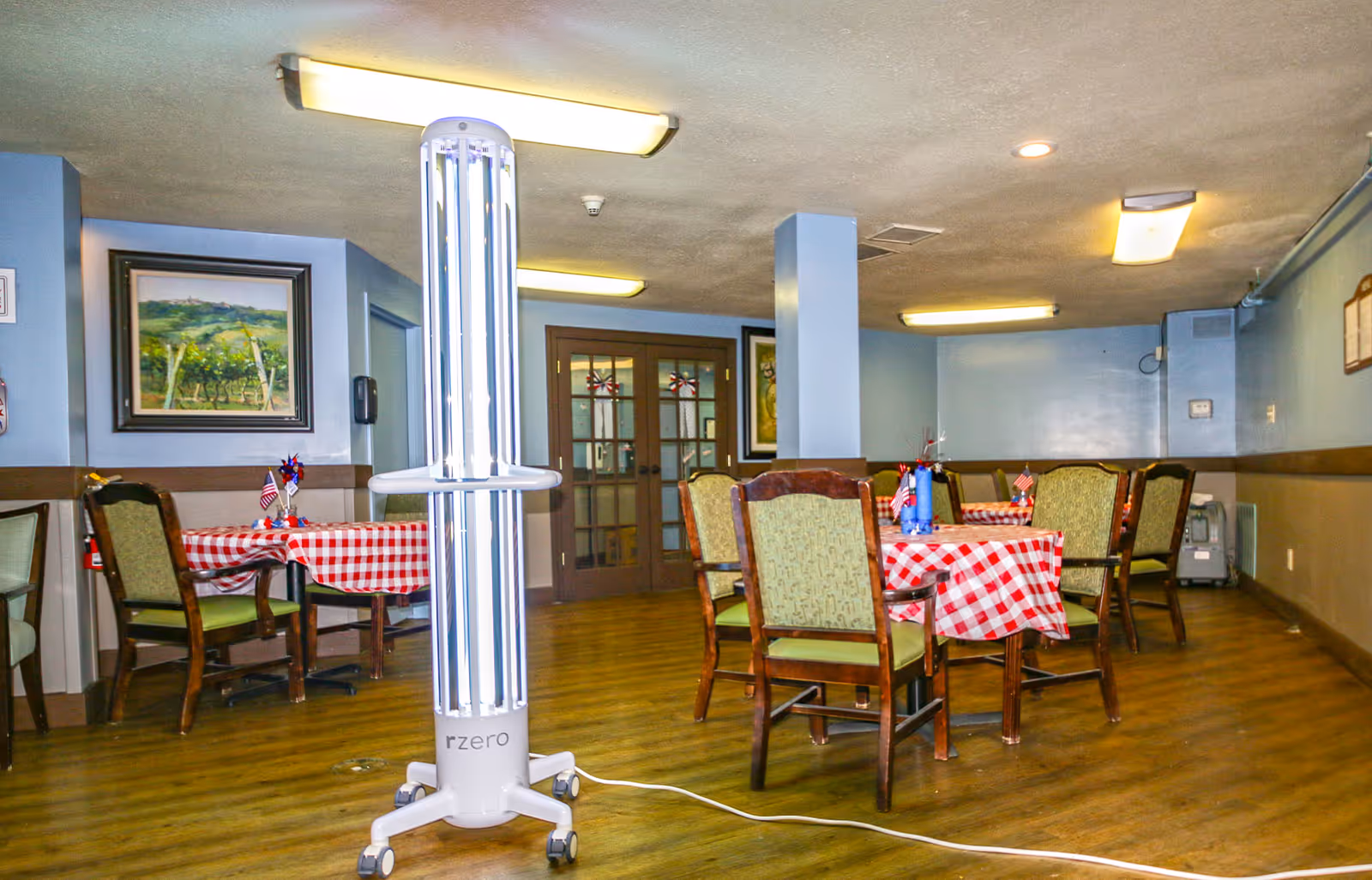 Dining room with wooden floor and several tables covered with red and white checkered tablecloths. Each table has chairs with green upholstery and small patriotic decorations. A tall white UV light sanitizer device on wheels is positioned in the center of the room. The walls are painted light blue with framed paintings and double glass doors in the background.