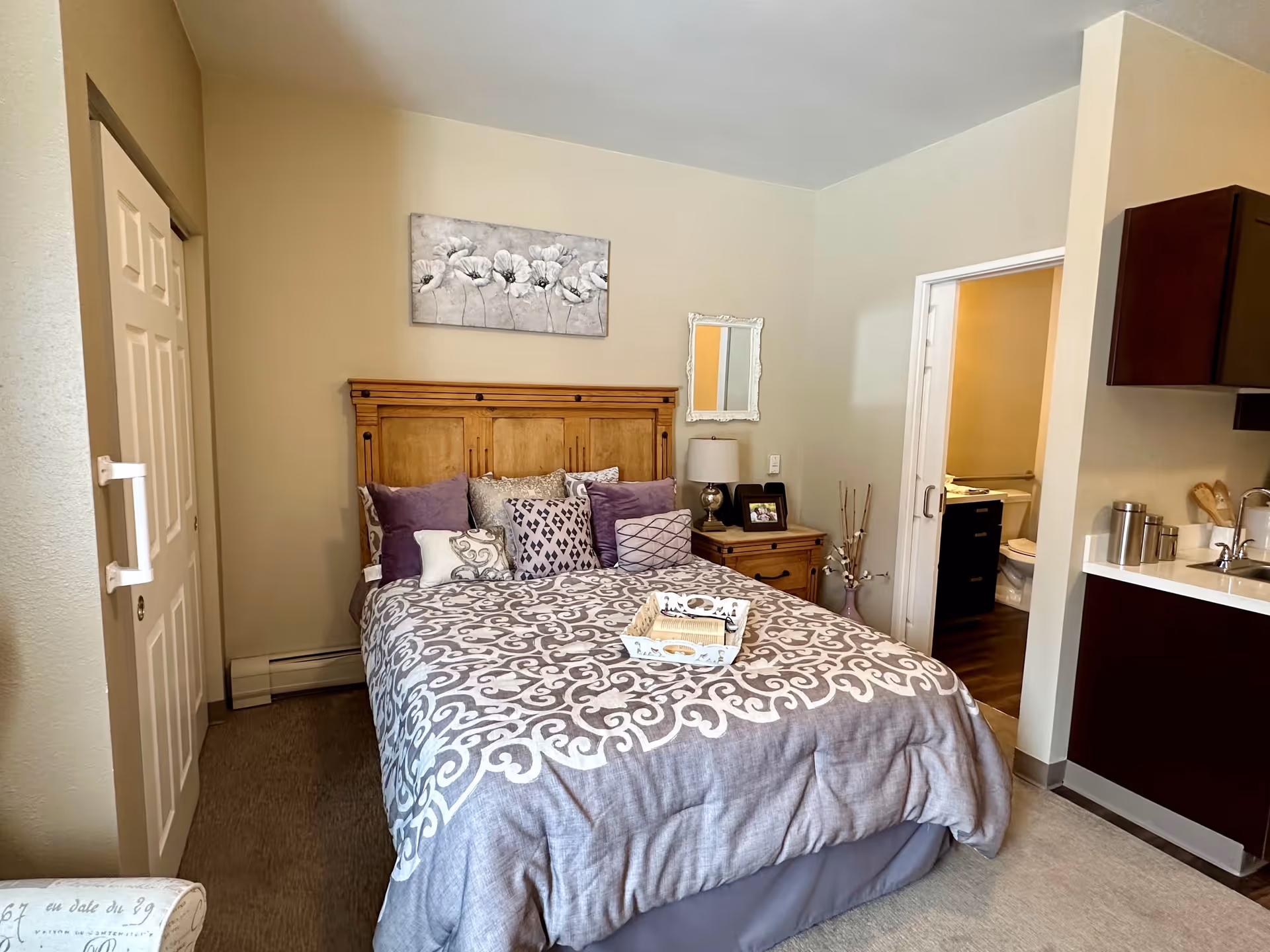 A cozy bedroom with a wooden headboard bed adorned with multiple decorative pillows and a patterned comforter. There is a nightstand with a lamp, a framed photo, and decorative sticks in a vase. A small mirror and floral artwork hang on the wall above the nightstand. To the right, there is a partial view of a kitchenette with a sink and cabinets, and an open door leading to a bathroom with a visible toilet.