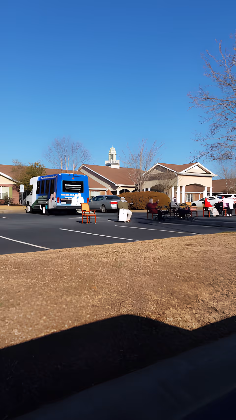 Parking lot outside a senior living facility with a blue and white shuttle bus, several parked cars, and a few chairs placed in the lot. The building has a beige exterior with a brown roof and a cupola on top. Leafless trees and a clear blue sky are visible in the background.