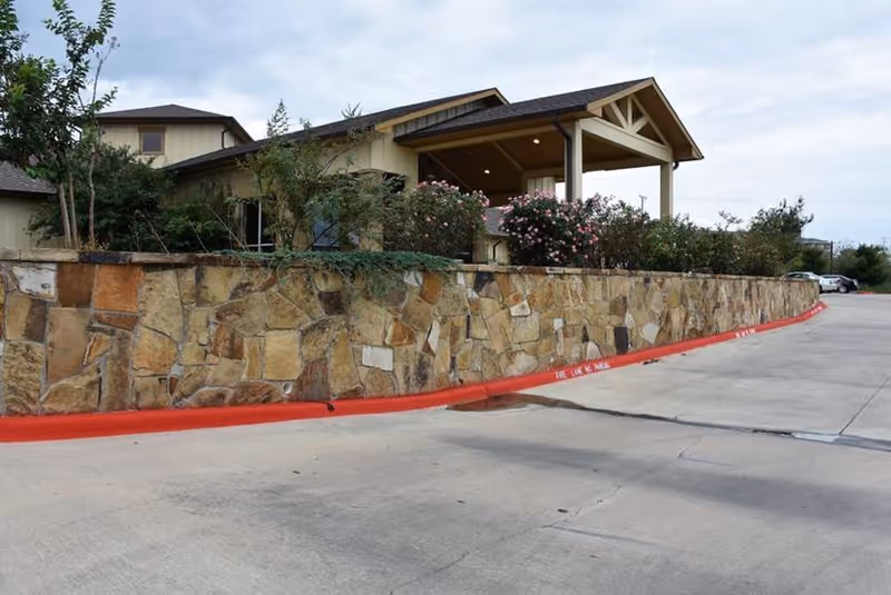 Exterior view of a building with a stone wall and a covered entrance. There are bushes and trees planted along the stone wall, and a concrete driveway with a red curb in front of the building. The sky is partly cloudy.