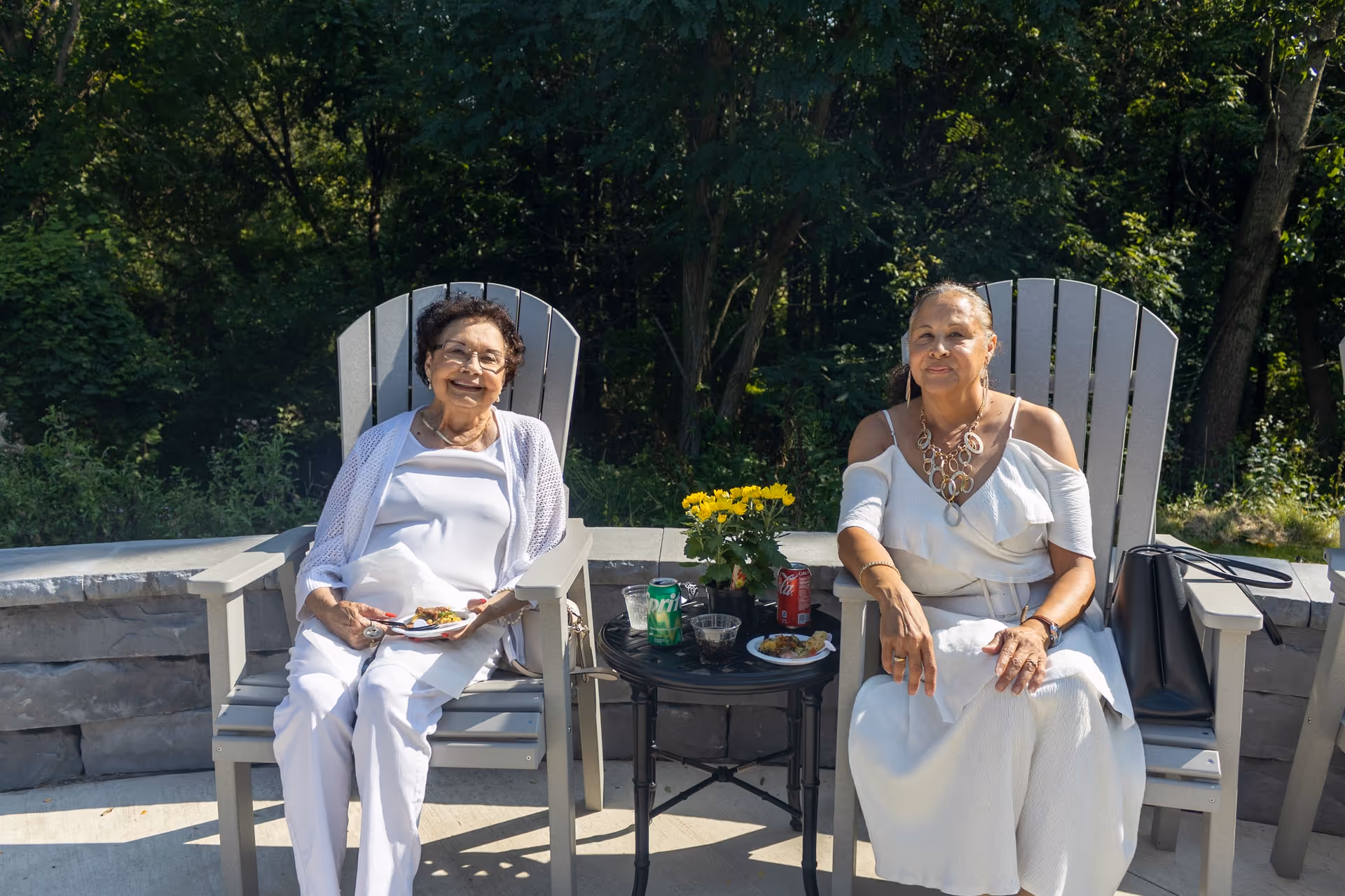 Two women sitting outdoors on gray Adirondack chairs with a small round table between them holding drinks, plates of food, and a yellow flower pot. They are surrounded by greenery and trees in the background, enjoying a sunny day.