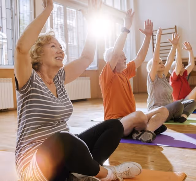 A group of elderly people sitting on yoga mats in a bright room, raising their arms above their heads as part of a seated exercise or stretching routine.