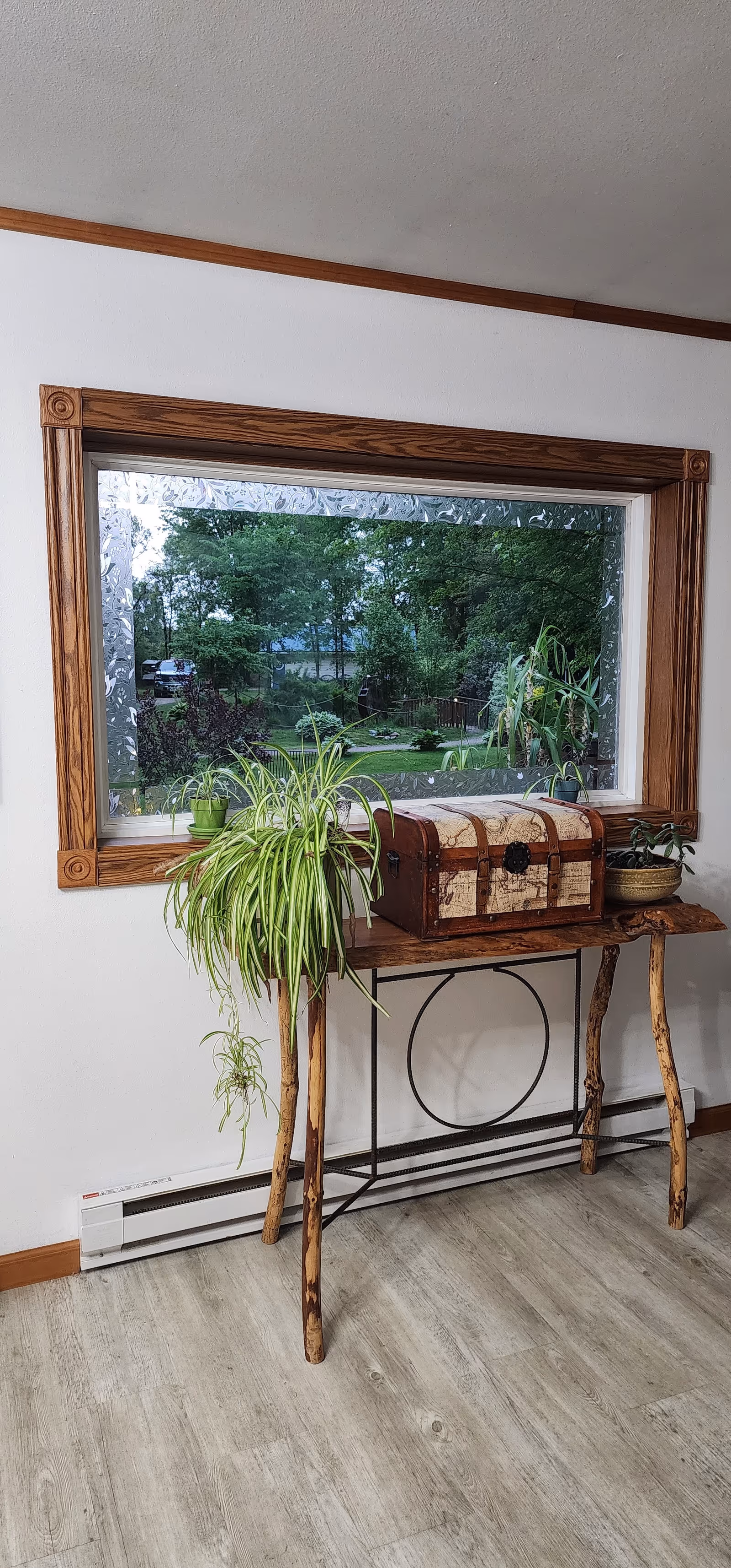 A wooden table with natural, rustic legs stands against a white wall beneath a large window with wooden trim. On the table are two potted plants and a decorative chest. Outside the window, green trees and a garden are visible.