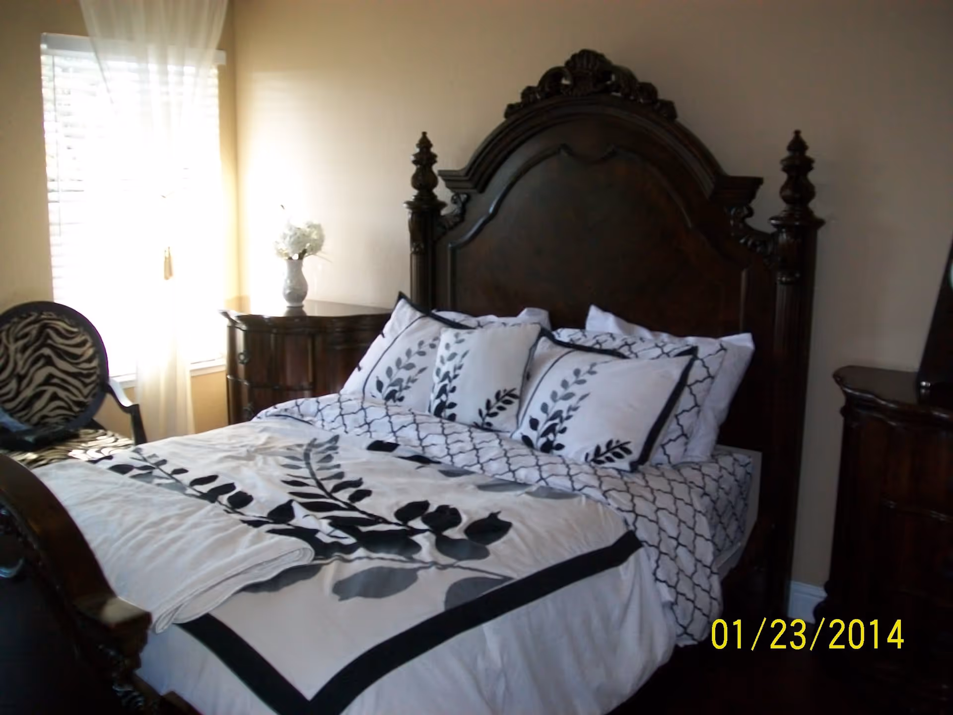 A bedroom with a large dark wooden bed frame featuring an ornate headboard. The bed is made with white and black bedding that has a leaf pattern, and there are several matching pillows. To the left of the bed is a window with sheer white curtains and a small wooden nightstand with a vase of white flowers. A chair with zebra-patterned upholstery is partially visible near the window. The walls are painted beige.