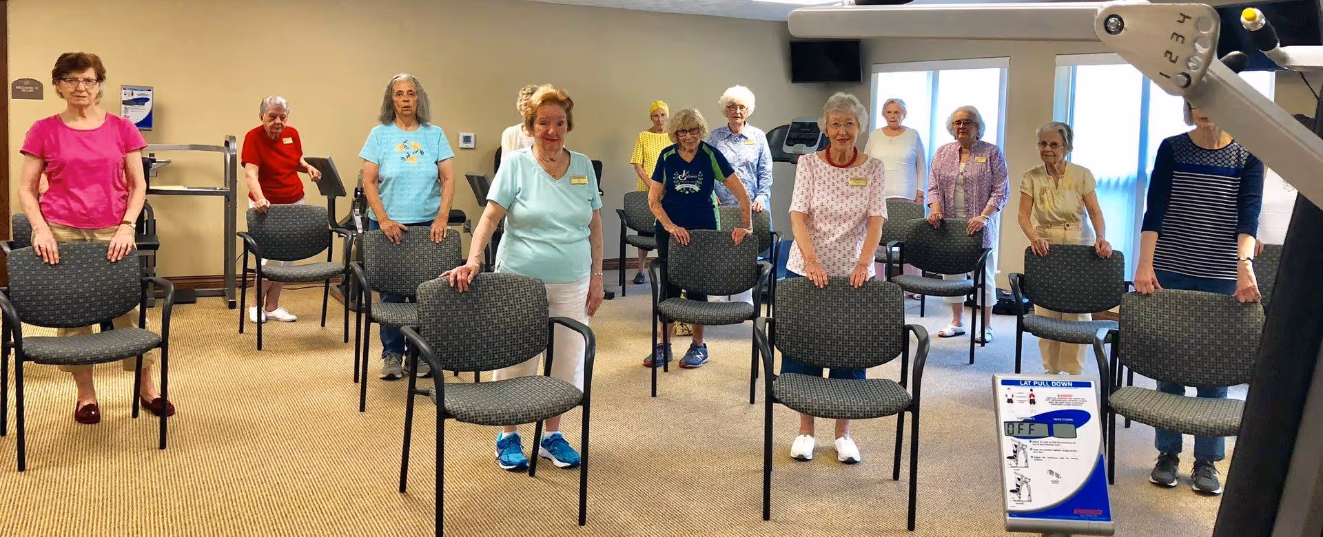 A group of elderly women standing behind chairs in a room, participating in a seated exercise or fitness class at a retirement facility.