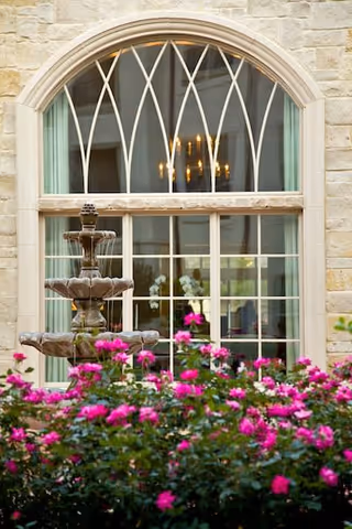 Stone exterior wall with a large arched window featuring decorative mullions. In front of the window, there is a tiered stone fountain surrounded by vibrant pink flowers and green foliage.